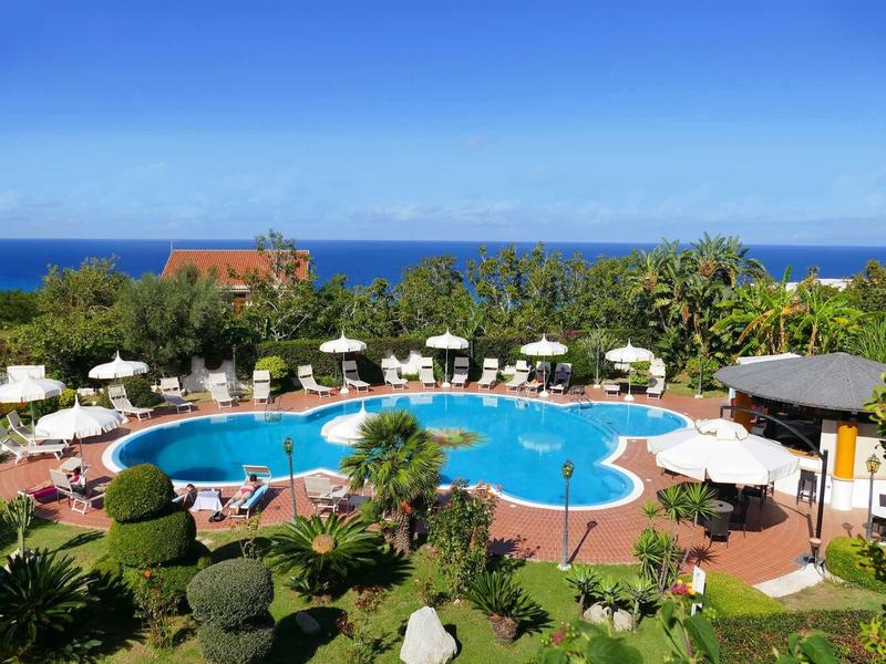 Pool in organischer Form, umgeben von grünen Pflanzen, weißen Sonnenschirmen und blauer Meereskulisse.