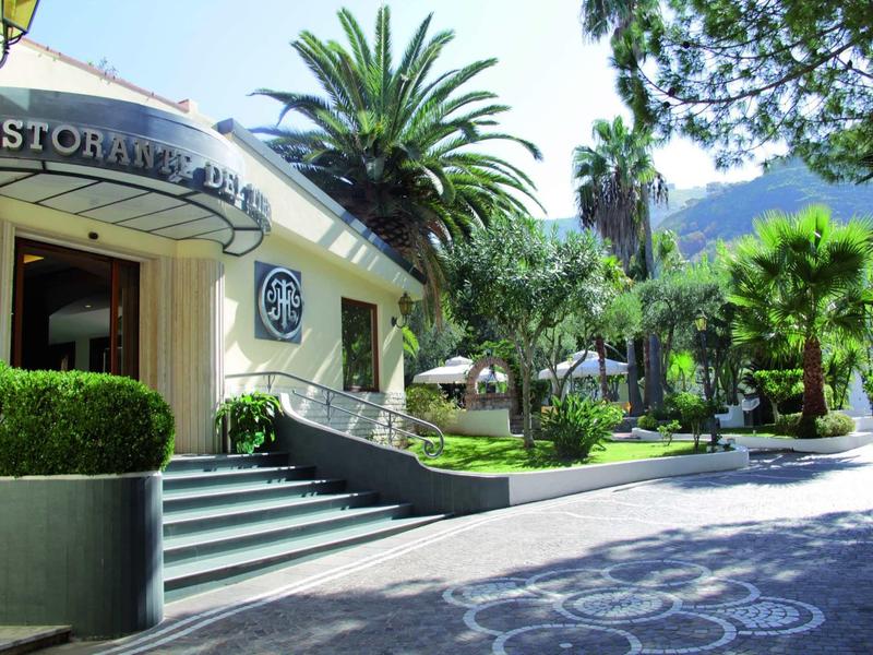 Eingang eines Gebäudes mit Treppe, Palmen und grüner Gartenanlage bei sonnigem Wetter.