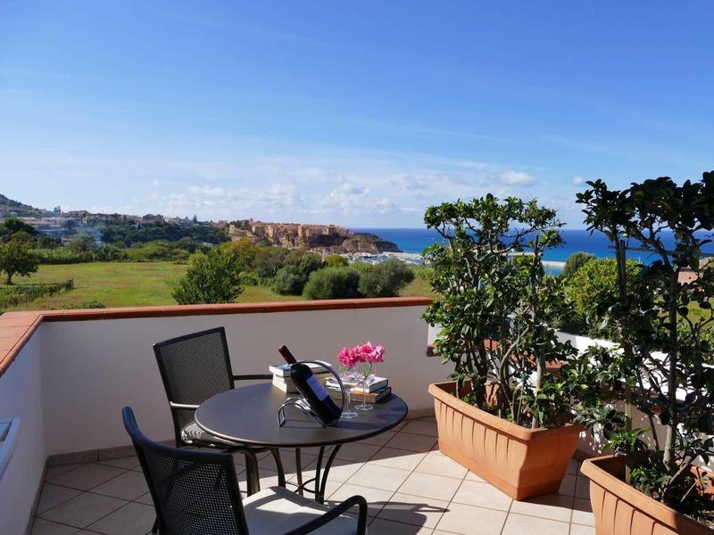 Balkon mit Tisch, Stühlen, Pflanzen in Töpfen, grünem Feld und blauem Meer im Hintergrund bei Sonnenschein.