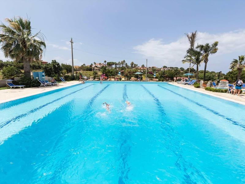 Une grande piscine extérieure avec une eau bleue claire et un espace bronzage entouré de palmiers.