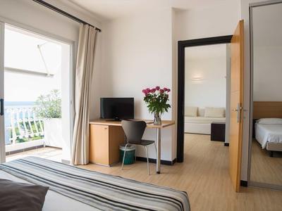 Chambre d'hôtel lumineuse avec balcon, bureau, chaise et fleurs à côté d'un cadre de porte ouvert.