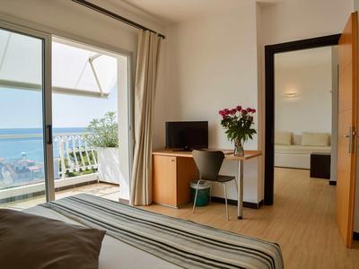 Chambre d'hôtel lumineuse avec vue sur la mer, balcon, bureau et fleurs sur la table.