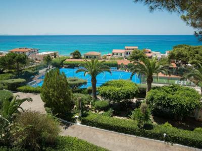 Vue sur un jardin d'hôtel avec piscine, palmiers et mer bleue en arrière-plan.