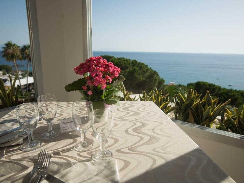 Table avec verrerie et bouquet de fleurs face à la mer et à la végétation côtière verte.