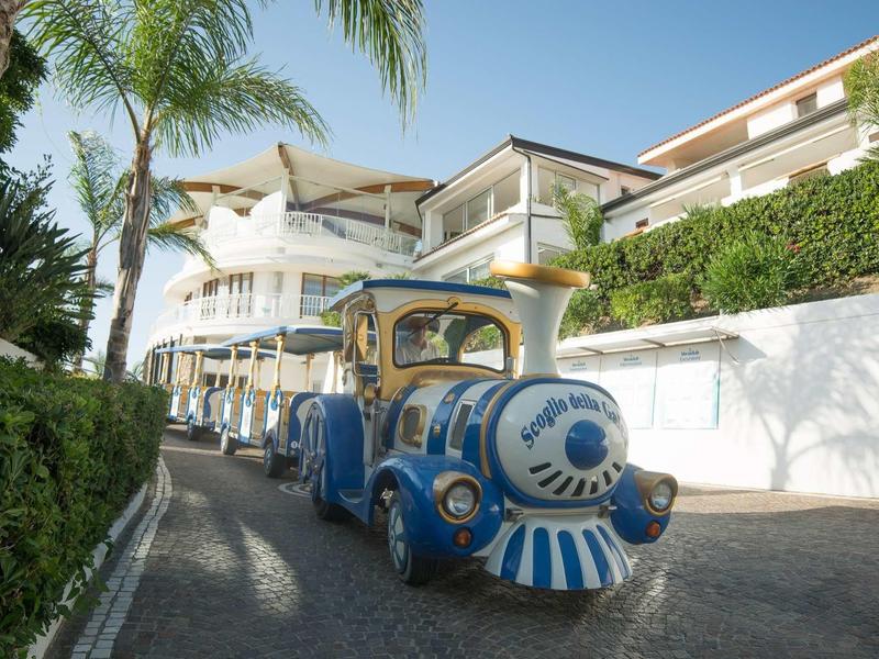 Petit train touristique bleu sur chemin pavé à côté d'un bâtiment blanc avec des palmiers.