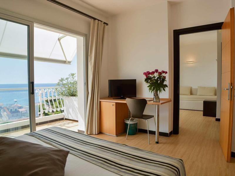 Chambre d'hôtel lumineuse avec vue sur la mer, balcon, bureau et fleurs sur la table.