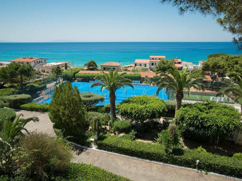 Vue sur un jardin d'hôtel avec piscine, palmiers et mer bleue en arrière-plan.