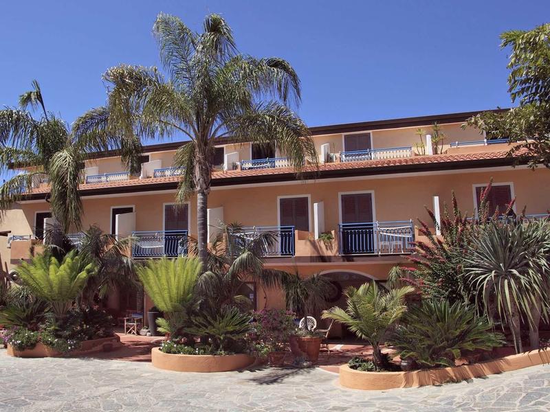 Zweistöckiges Haus mit braunen Wänden, mehreren Palmen und Sträuchern im Vorgarten bei blauem Himmel.