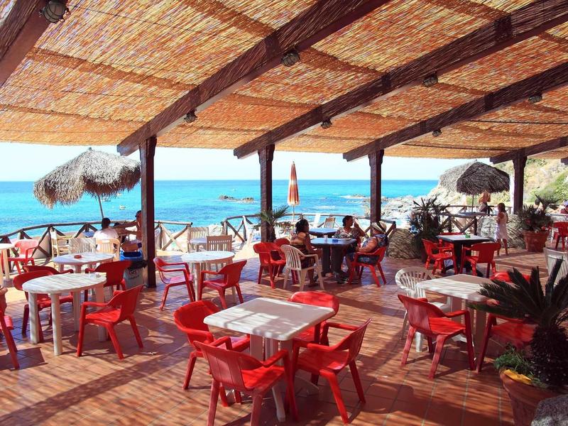 Strandrestaurant mit roten Stühlen, weißen Tischen, Holzdach und Meerblick im Hintergrund.