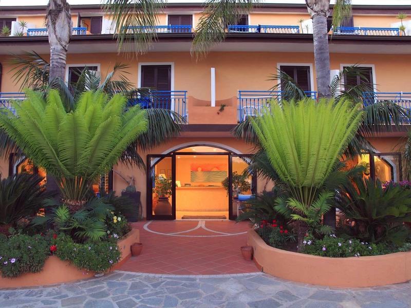 Eingang zu einem orangen Gebäude mit symmetrischen Pflanzbeeten und Palmen vor dem Tor.