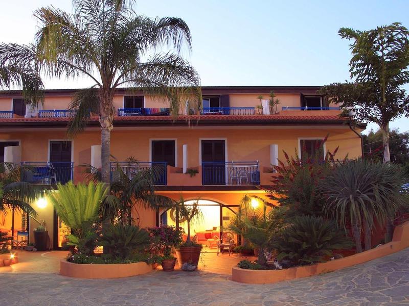 Mehrstöckiges Haus mit orangener Fassade, Palmen und Pflanzen vor Eingangsbereich bei Dämmerung.