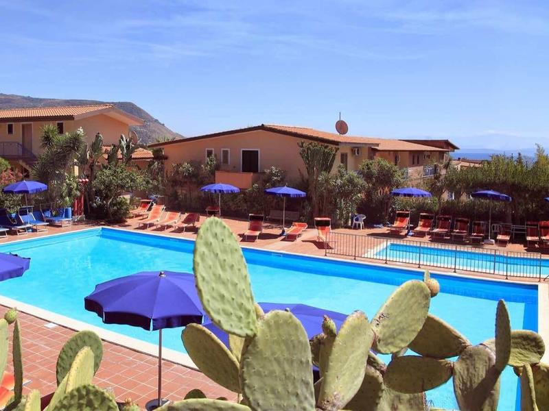 Blick auf einen großen Hotelpool mit blauen Liegen und kaktusähnlichen Pflanzen im Vordergrund.