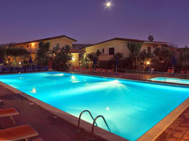 Beleuchteter Swimmingpool am Abend, umgeben von Sonnenliegen und niedrigen Häusern, klarer Himmel.