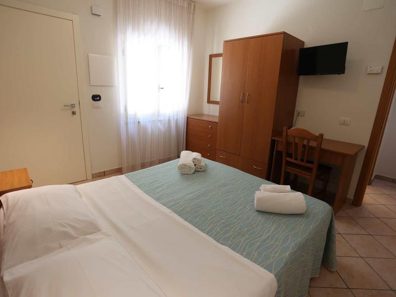 A simple hotel room with a double bed, desk, wardrobe, and wall-mounted TV.
