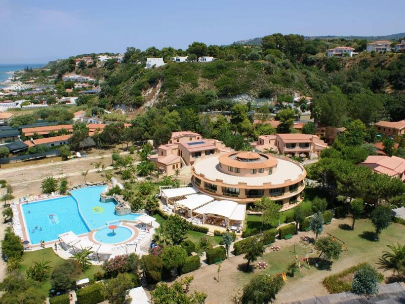 Aerial view of a hotel with blue pool near coast and green hills in the background.