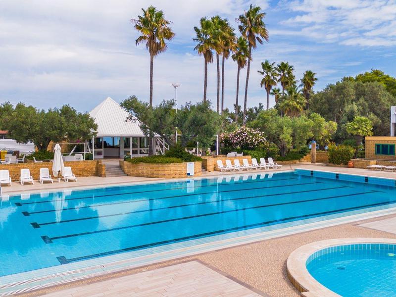 Piscine extérieure avec chaises longues, parasols et palmiers dans un hôtel sous un ciel bleu.