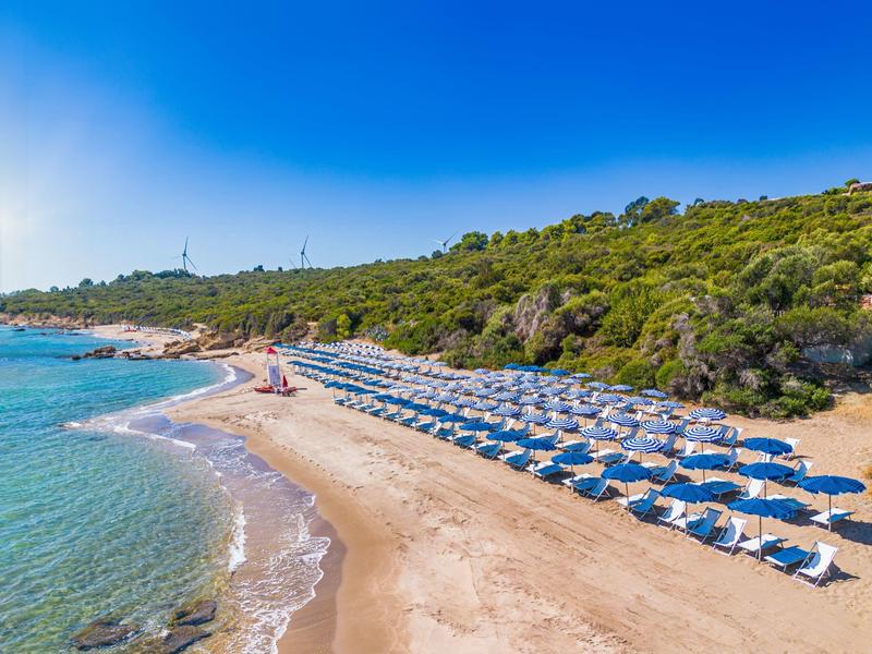 Plage avec parasols bleus et mer claire devant une colline boisée sous un ciel bleu.
