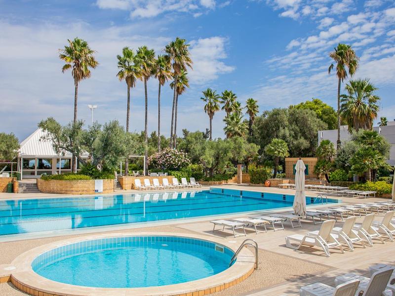Piscine d'hôtel avec chaises longues et palmiers sous un ciel bleu.