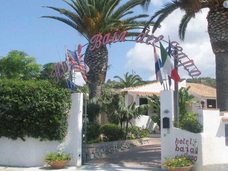 Entrée avec palmiers et drapeaux vers un hôtel sous un ciel clair et ensoleillé.