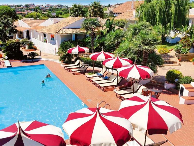 Une piscine extérieure ensoleillée avec des parasols rouges et blancs à côté de bâtiments méditerranéens.