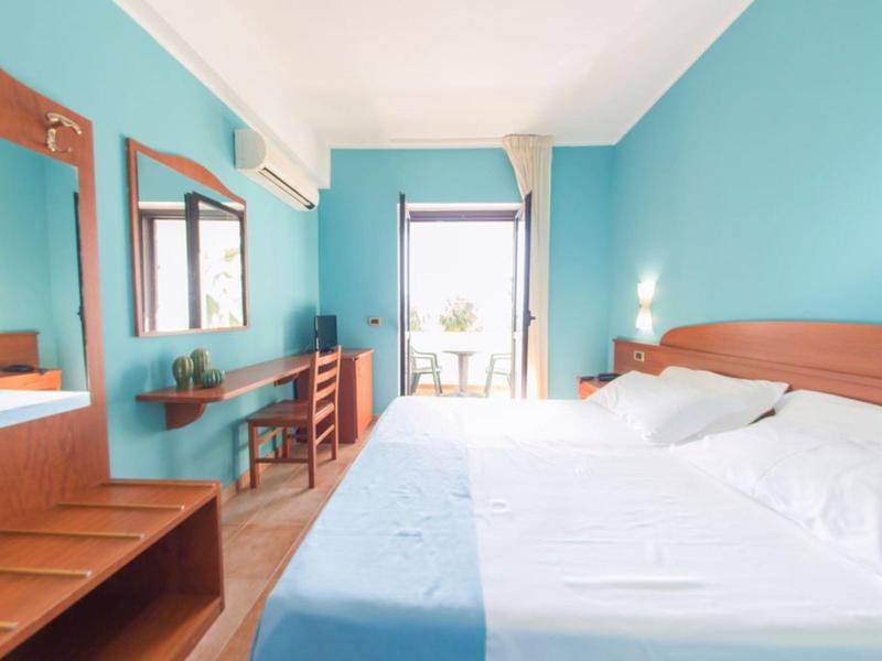 Chambre d'hôtel lumineuse avec grand lit, murs bleus et balcon avec vue sur l'extérieur.