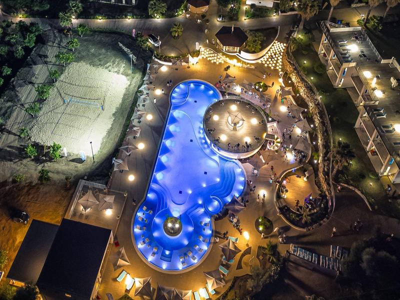 Vue nocturne d'une piscine éclairée dans un complexe hôtelier avec des bâtiments et plantations.