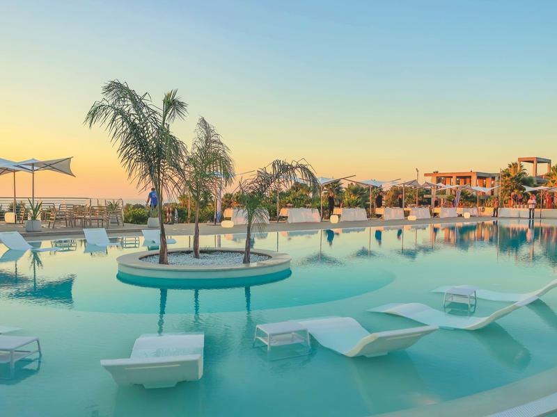 Piscine avec chaises longues immergées, palmiers et coucher de soleil dans un complexe hôtelier.