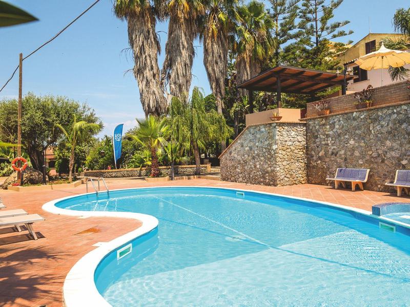 Piscine extérieure curviligne entourée de palmiers et de chaises longues sous un ciel bleu.