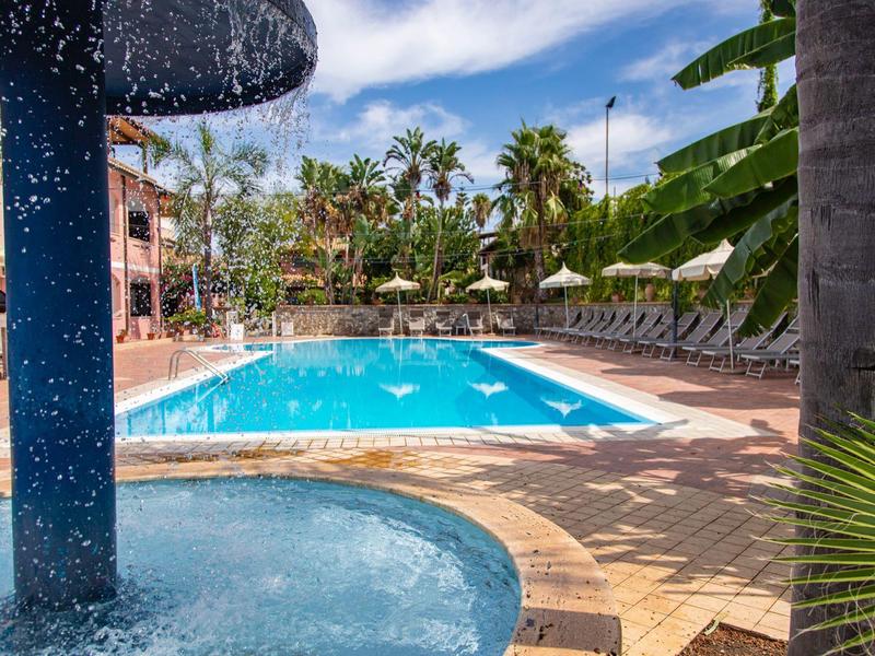 Piscine d'hôtel avec cascade, chaises longues et palmiers sous un ciel partiellement nuageux.
