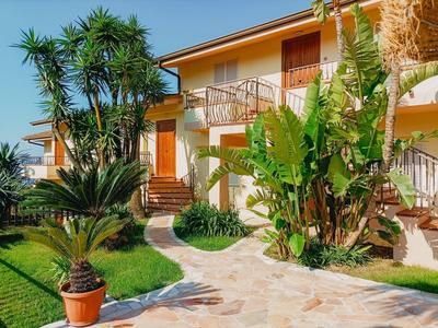 Chemin serpentant entre plantes tropicales vers une maison de vacances jaune à deux étages sous un ciel bleu.