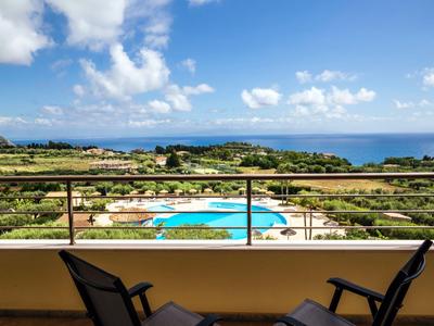 Uitzicht vanaf balkon met stoelen op zwembad, tuin en zee onder een blauwe hemel met wolken.