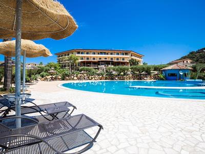 Hôtel avec grande piscine, transats et parasols en paille sous un ciel bleu clair.