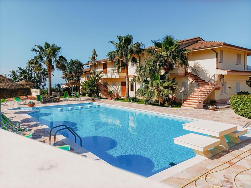 Un hôtel avec une grande piscine extérieure, entourée de palmiers et de chaises longues sous un ciel bleu.
