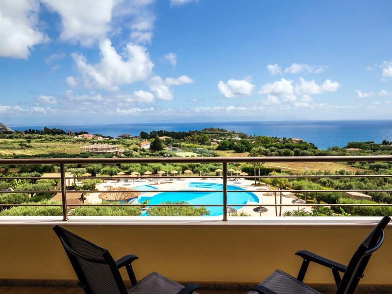 Vue depuis un balcon avec des chaises sur une piscine et la mer sous un ciel bleu avec des nuages.