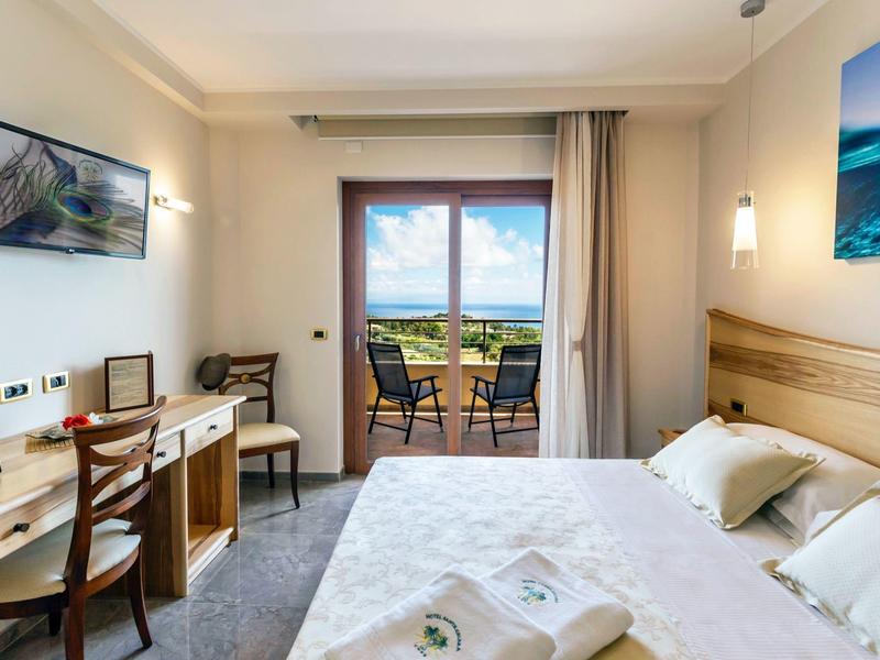 Chambre d'hôtel lumineuse avec lit double, bureau, télévision et balcon donnant sur un paysage verdoyant.