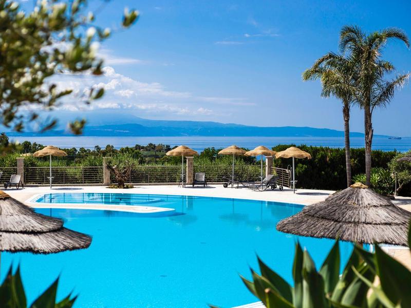 Grande piscine avec parasols en paille et vue sur la mer sous un ciel clair.