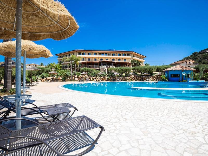 Hôtel avec grande piscine, transats et parasols en paille sous un ciel bleu clair.