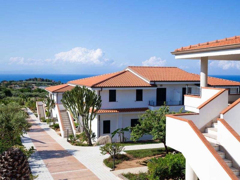 Nowoczesny kompleks hotelowy z czerwonymi dachami i widokiem na morze pod niebieskim niebem.