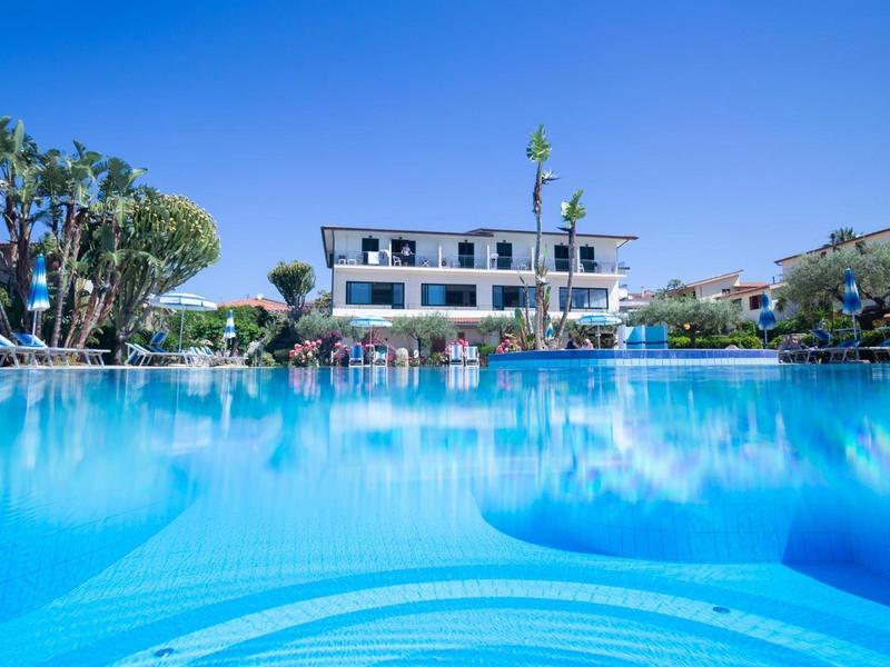 Duży niebieski basen przed wielopiętrowym hotelem pod bezchmurnym niebem.