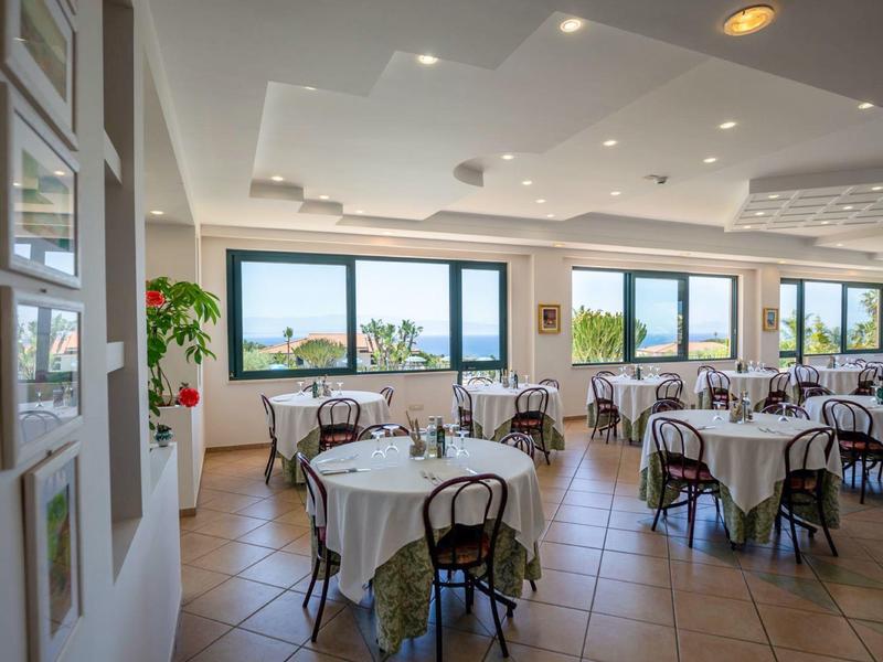 Jasna restauracja z okrągłymi stołami, widokiem na morze i klify w świetle dziennym.