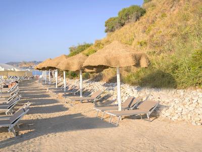 Row of empty sun loungers with straw canopies on sandy beach by a leafy hillside.