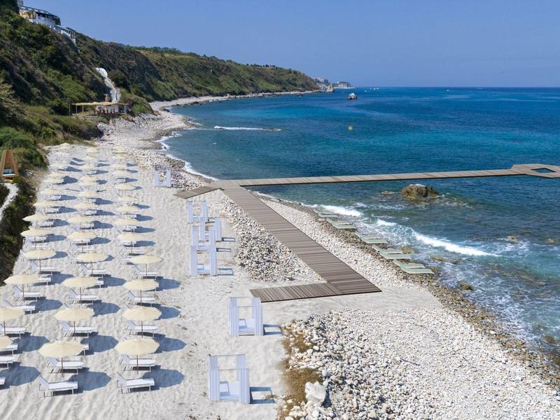 Une plage de galets avec des parasols blancs et des chaises longues au bord de la mer bleue sous un ciel clair.