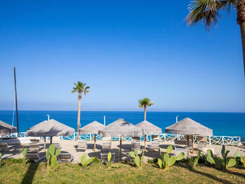 Strandresort met ligstoelen en rieten parasols tegen een heldere blauwe lucht en zee.
