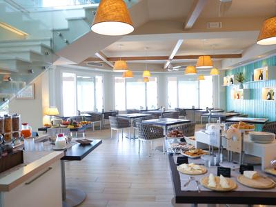 Helles, modernes Café mit großen Fenstern, Sitzbereichen, Buffet und Holz- sowie weißen Farbtönen.