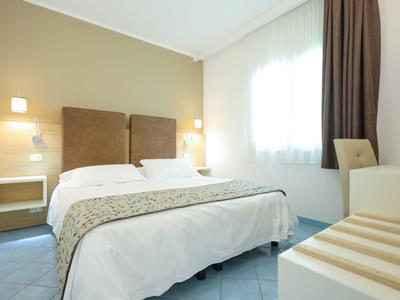 Bright hotel room with a double bed, bedside lights, a window with curtains, and a chair.