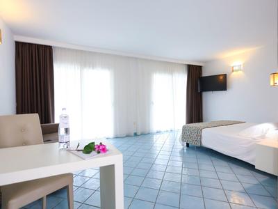 Bright hotel room with tiled floor, bed, table with chair, and large window with curtains.