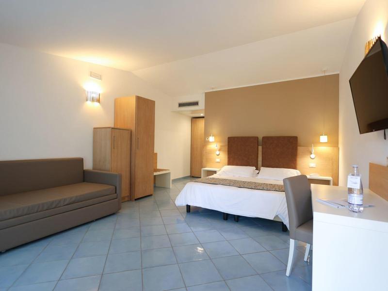Modern hotel room with double bed, sofa, TV, desk, and tiled floor in beige and brown tones.