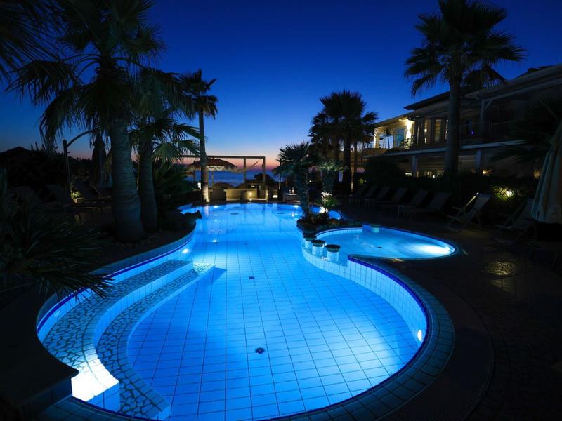 Outdoor pool at twilight with glowing blue water and surrounding palm trees and building lights.