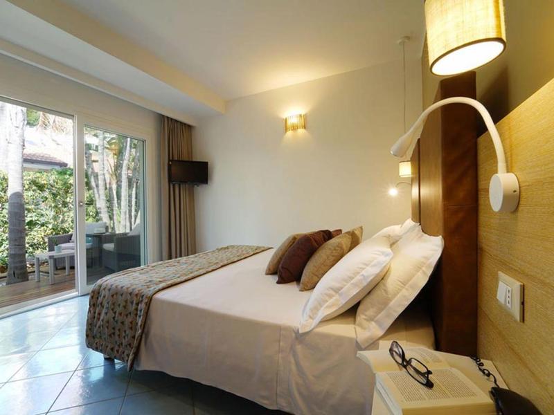 Hotel room with double bed, bedside lamps, and glass doors opening to a garden.