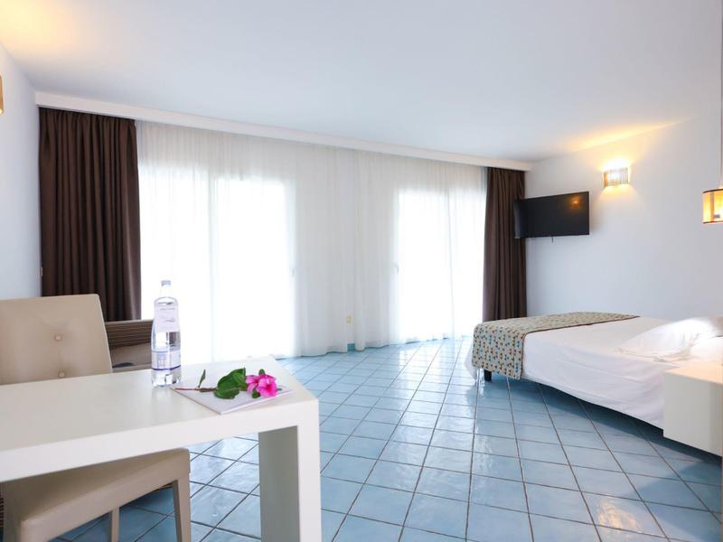 Bright hotel room with tiled floor, bed, table with chair, and large window with curtains.